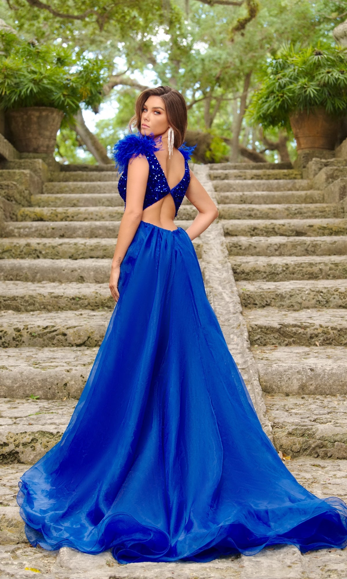 A woman stands on stone steps in an outdoor garden, wearing the Long Formal Dress 38339 by Ava Presley—a royal blue, v-neck evening gown with feathered shoulder straps—glancing over her shoulder.